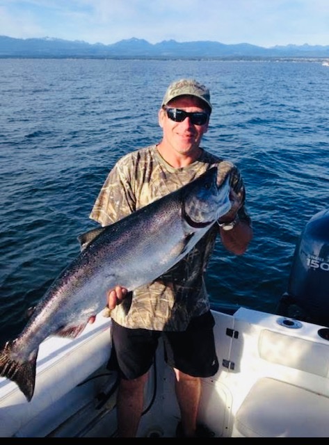 Douglas holding a fish he caught, on a boat. Calm waters, mountains and cloudy skies are visible in the background behind him.